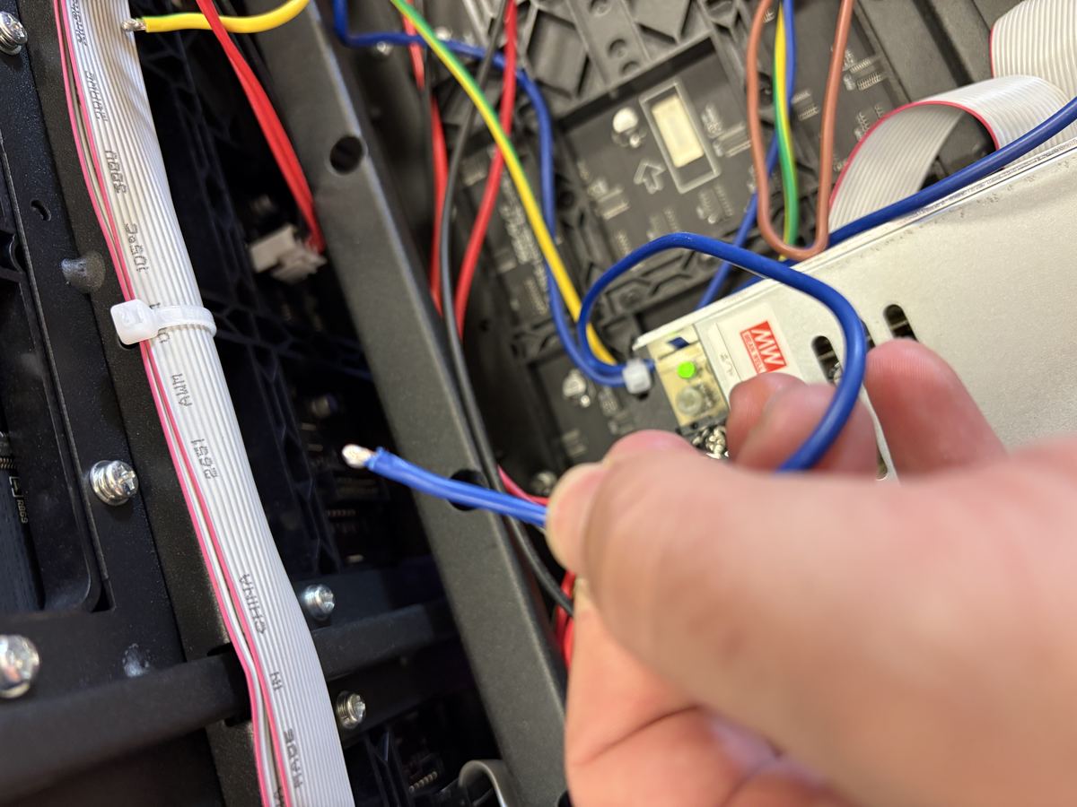 Hand grasping stripped blue mains conductor — green LED on PSU confirms unit is energised